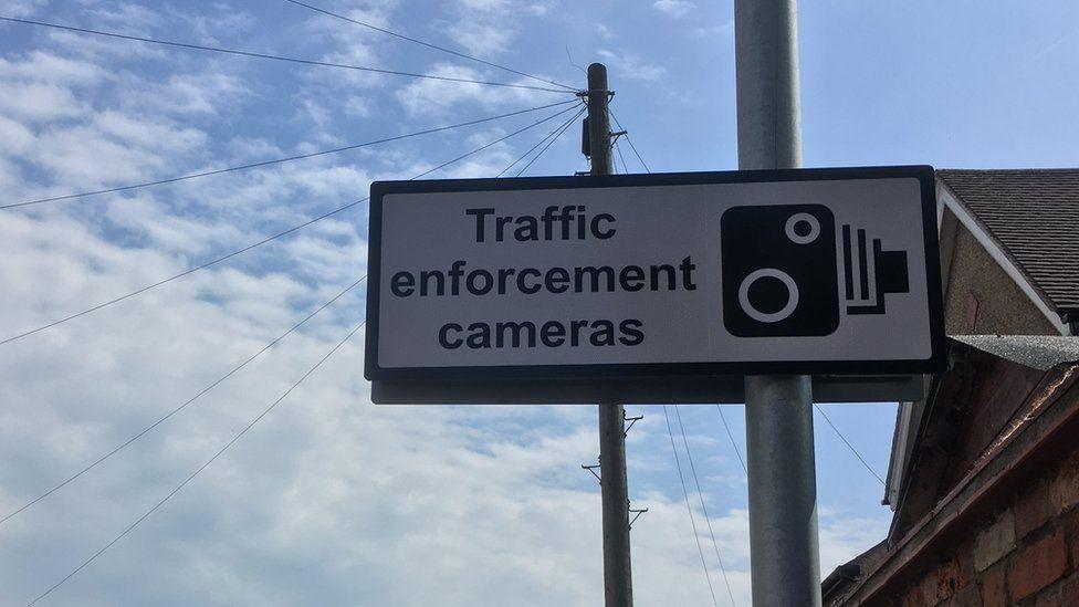 White sign on a pole, with black lettering "Traffic Enforcement Cameras" and a camera symbol. A house roof and the sky are visible behind.