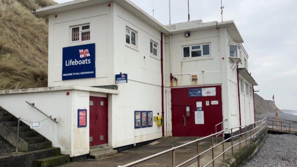 The lifeboat station in Sheringham. The building is white and has two levels with red doors. There is a sign on the side of the building which is a blue rectangle with the RNLI logo on it. Behind the lifeboat station are cliffs.
