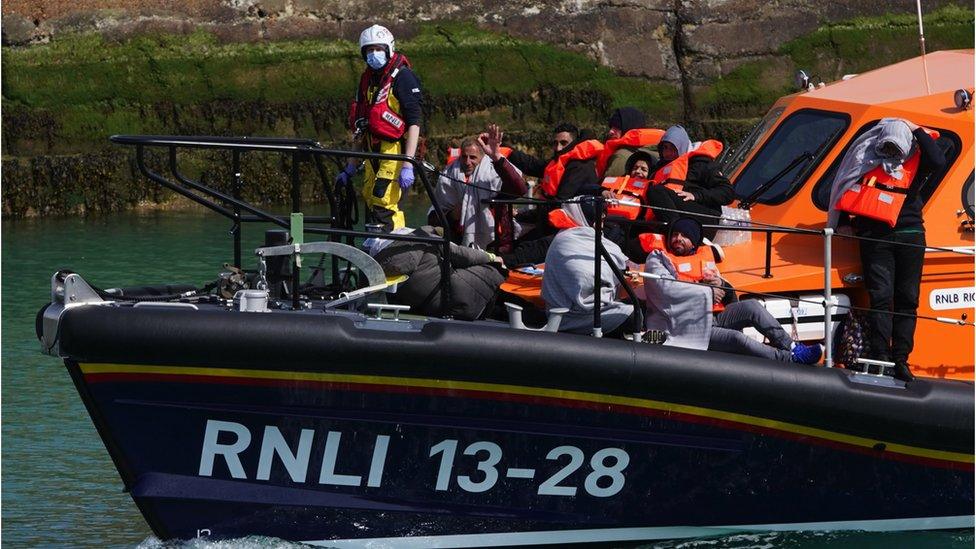 A lifeboat brings migrants into a harbour, men in lifejackets seen waving from the deck