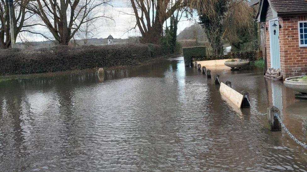 A flooded road with a cottage at the side with water at the door and sandbags