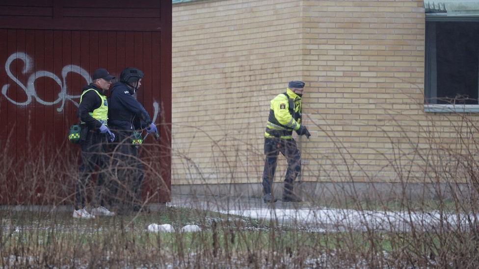 Three police officers with guns standing around the side of a building