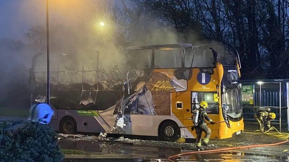 A yellow bus which has been badly burnt by a fire. The fire has particularly affected the top of the bus toward its back. Two firefighters are working near the front of the bus.