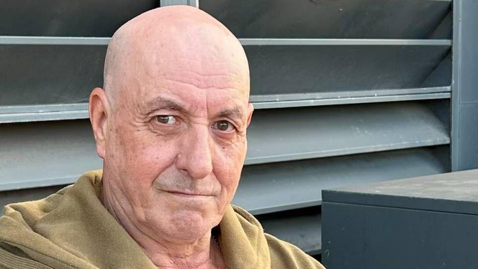 A man with no hair, wearing a yellowish hoody is looking at the camera