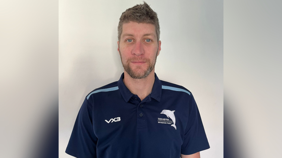 A man wearing a sports top with the Tadcaster Community Swimming Pool logo on looks at the camera and smiles.