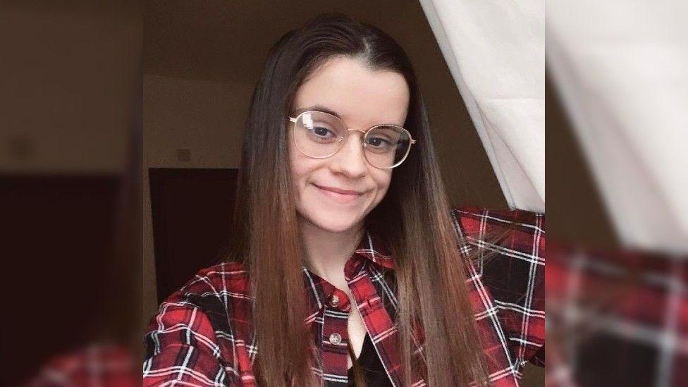 A young woman with long hair and glasses, wearing a red checked shirt.