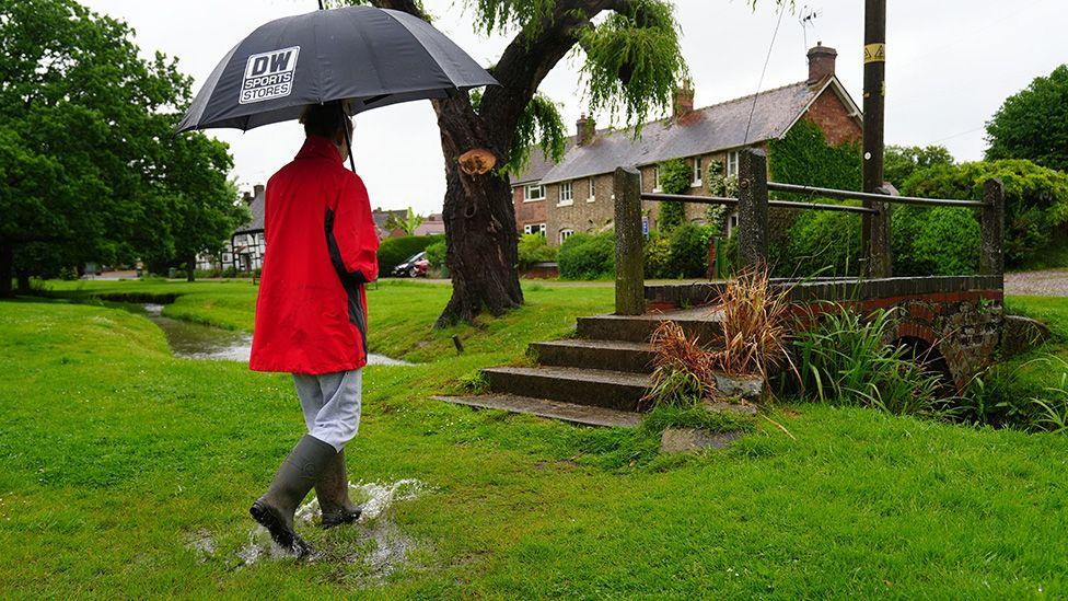 A person walks with an umbrella in a waterlogged area of grass in front of a foot bridge in the village of Charlton on 22 May