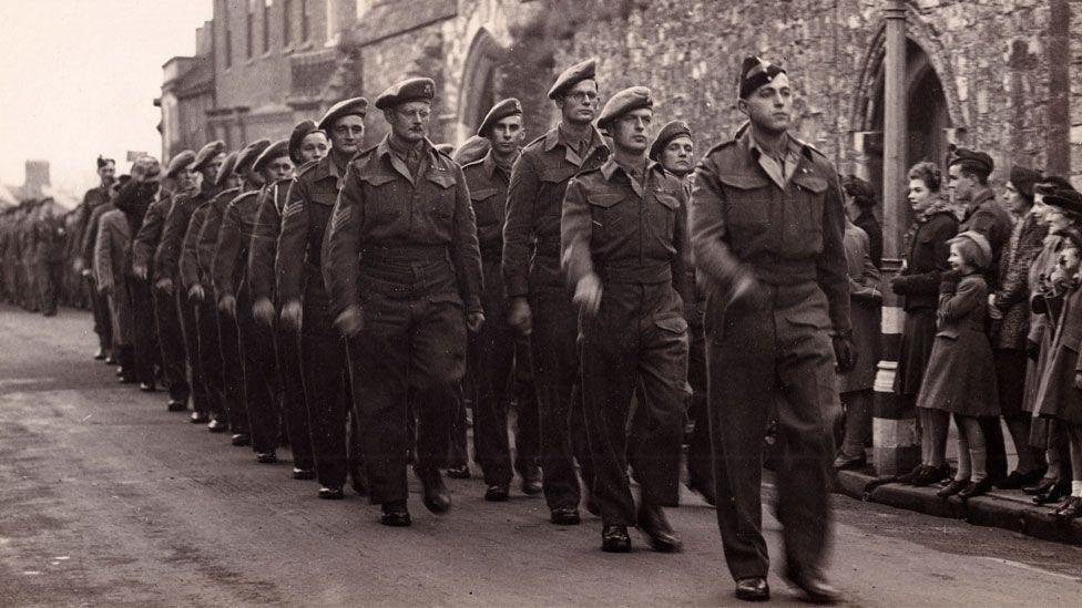 A black and white image showing men from the Cambridgeshire Regiment marching past Ely's war memorial. They are in Army uniform. They are being watched by men, women and children.