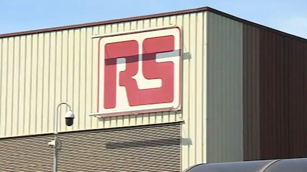 Part of a cream metal building with a large RS logo fixed to its side.