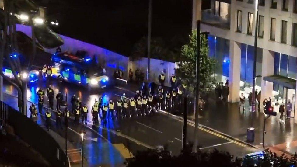 A line of more than 30 police officers moves towards crowds in Stratford, with two police vans following behind with their blue lights on.