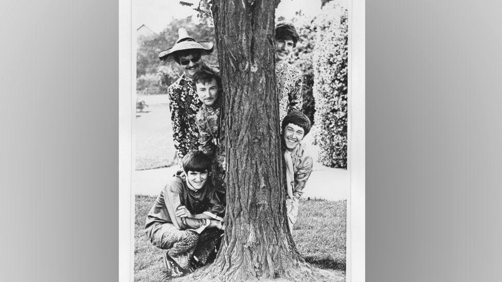 A black and white photo of the band posing in Memorial Park, Luton. The photo is portrait. The lower half of a tree trunk is in the centre, with three men leaning out on one side and two on the other. Chris wears sunglasses and a pointed Mexican style straw hat. There are landscaped hedges and lawns behind them.