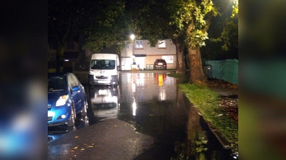 A residential road with water covering it in Cheltenham at night. There is a blue car and a van parked on the side of the road.