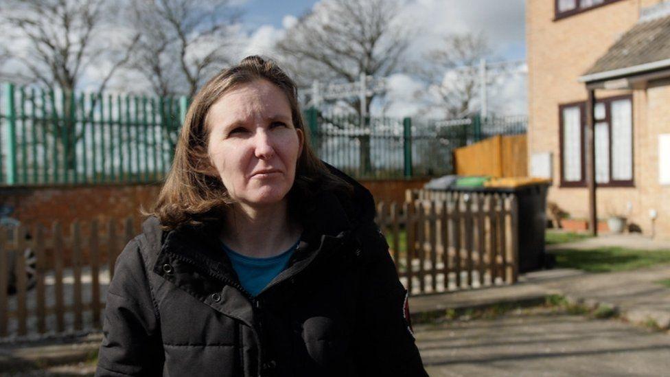 Julia Virdee standing outside, by a railway line and homes. She is looking away, wearing a dark coat with a blue top underneath. She looks concerned. You can see trees in the back ground, without leaves on, and railings. 