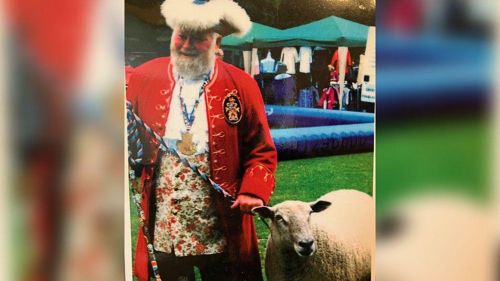 A picture of Trevor Heeks in his full town crier outfit, including a large fur-lined hat and red jacket, pictured walking a sheep. 