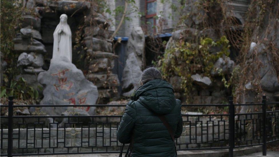 Woman, wrapped in a winter coat, prays in front of a statue of the Virgin Mary.