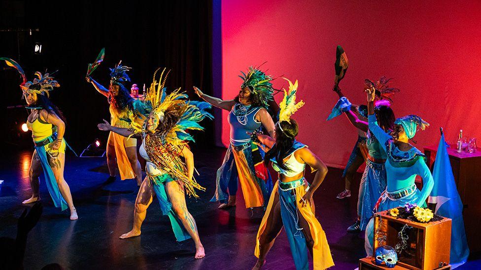 Dancers perform on stage wearing brightly coloured yellow ad blue costumes. 