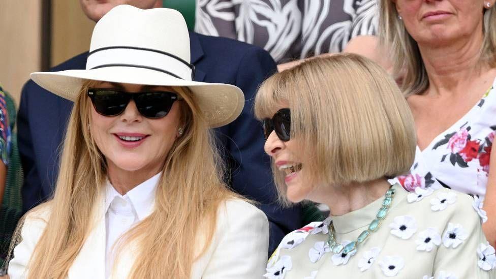 Nicole Kidman and Anna Wintour smile as they sit next to one another at Wimbledon. Kidman is wearing a white sun hat and sunglasses, and Wintour is wearing a flowery dress with her tradition blunt, blonde bob.