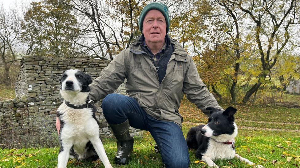 A man in a green hat and green waterproof coat and blue jeans is kneeling down with a sheepdog on either side of him. There is a dry stone wall behind him and trees in the background.