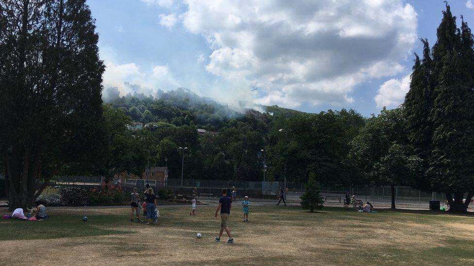 Cloud of smoke from a fire with people in a park looking at it from below. A helicopter is in the smoke cloud