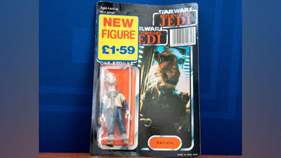 A Yak Face toy figurine, with a price tag on, showing a cream figure, wearing blue trousers and top, and an image of him from the Star Wars film, next to the figurine.