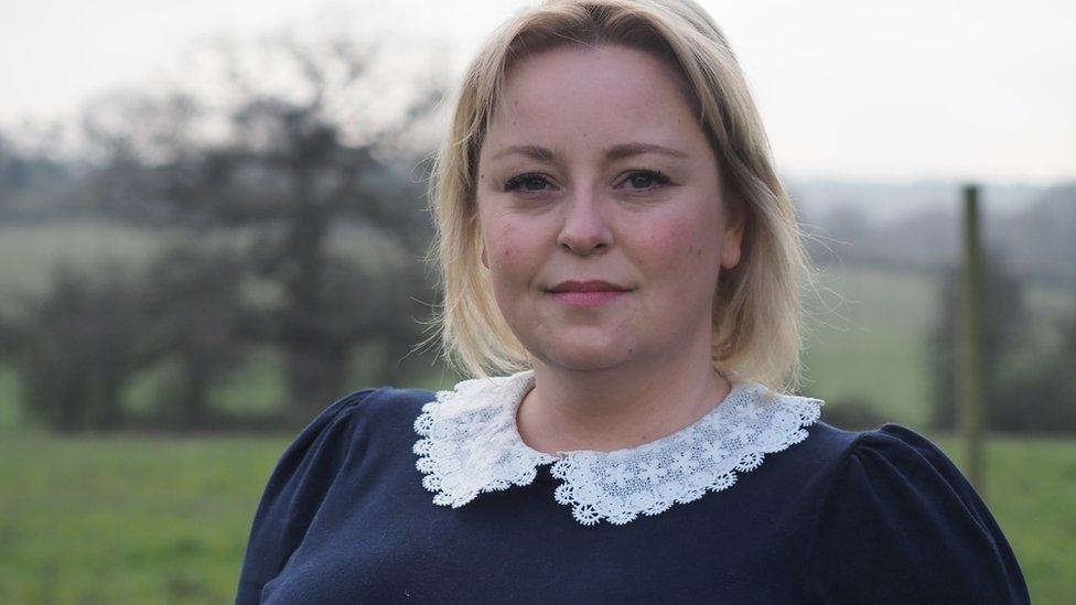 A white middle aged woman with shoulder length blonde hair wears a navy jumper with a white lace collar and looks at the camera