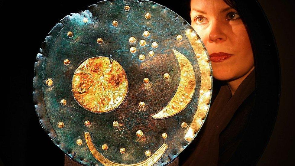 a woman poses with a bronze disc covered with a sun and a moon and lots of stars