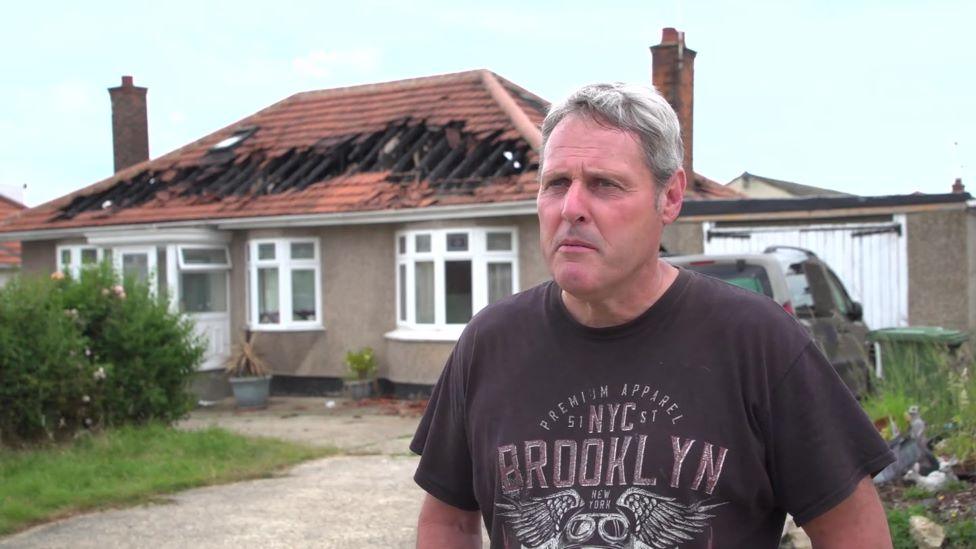 Neighbour David Bridge stands in front of the fire struck home. He is wearing a dark coloured t-shirt and behind him you can see the burnt out remains of the roof.