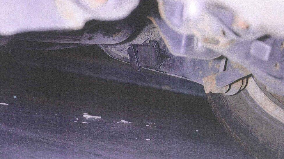 Close up image of a small, square black tracking device fitted under a Skoda car used by Stephen "Bonzo" Daniel. One of the wheels is visible in the image and some rust is visible.