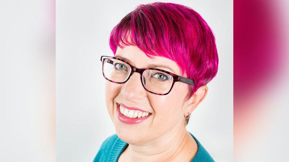 Brenda Coombes is smiling at the camera and wearing a green jumper and has pink hair