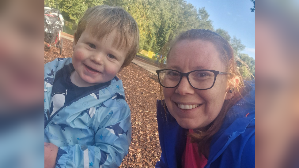 Emma Street and her young son Connor smile at the camera, he has dark blonde hair and red cheeks. He is wearing a waterproof jacket with cartoon sharks on it. Emma wears black glasses and smiles at the camera, she wears a blue waterproof jacket. They appear to be in a playground with bark on the floor