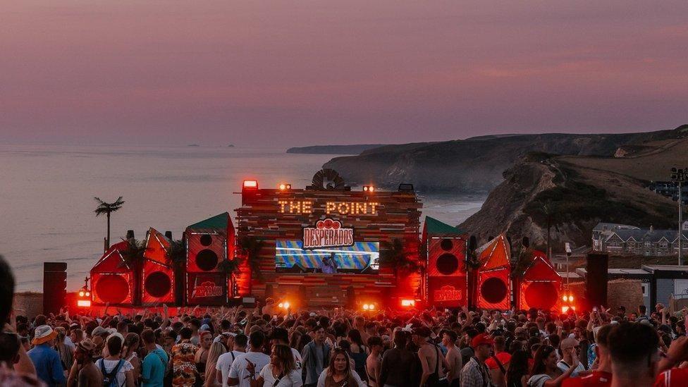 The festival with a stage which says The Point in lights on it, there are lots of people gathered and some speakers either side of the stage. The sea is also behind it.