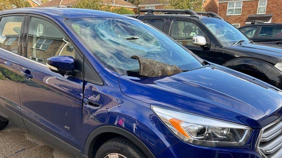 Bright blue car with windscreens smashed and brown roof tile wedged by wiper blades