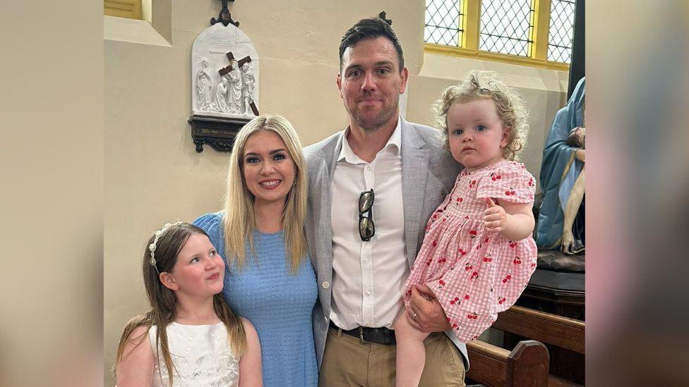 Glen stands with his wife and two children in a church. They are dressed as wedding guests, and are smiling at the camera. Their oldest daughter is looking and smiling at Amber.