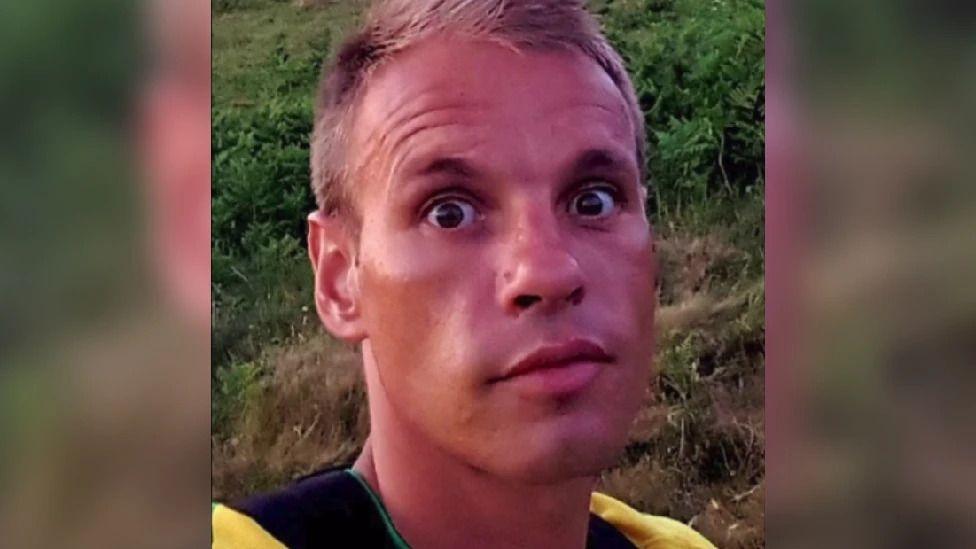 Jordan Moray looks at the camera, he is wearing a yellow top and shrubs and grass is visible behind him.