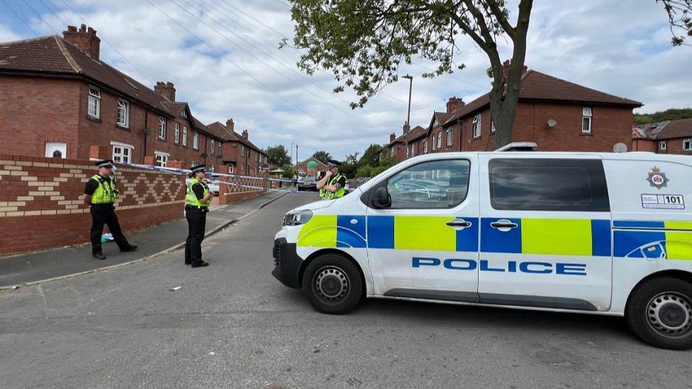 A police van is parked on a road in a residential street, with three police officers wearing their uniforms and hi-vis jackets stood on the road too. A police cordon is in place in the background.