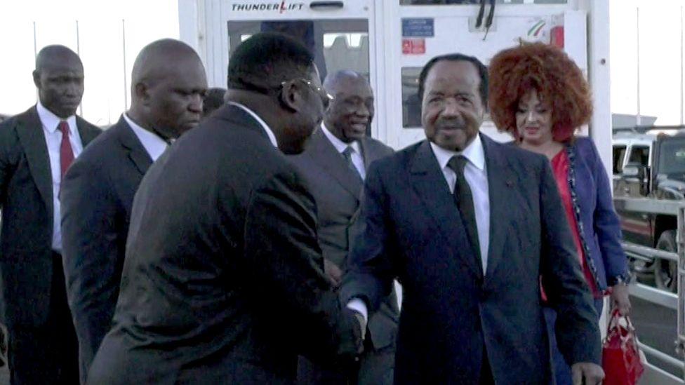 Paul Biya being greeted by an official at the airport. His wife, Chantal, can be seen in the background.