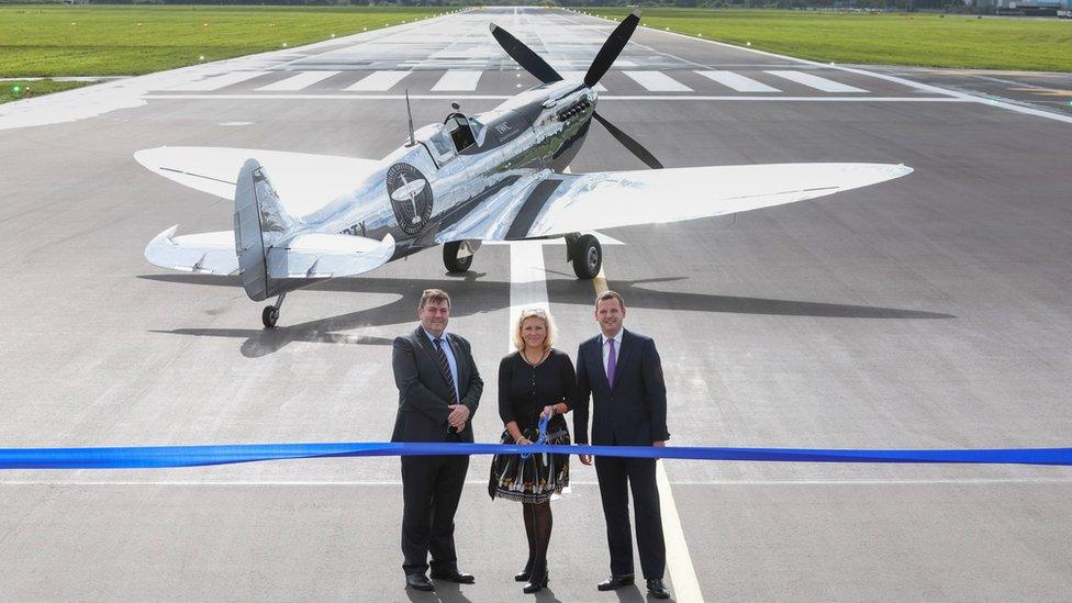 Three people stand in front of a runway about to cut a blue ceremonial ribbon