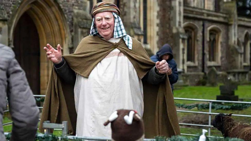 A man dressed as a shepherd in a live nativity scene near the cathedral. He has a brown cloak, a piece of head-wear with a tea-towel hanging around his head, and is greeting people who are off-camera. Next to him is a black sheep.