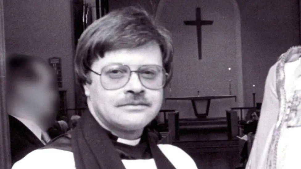 A vicar dressed in vestments and wearing 1980s-style glasses looks into the camera against the backdrop of a church altar and a cross above it. The image is monochrome.