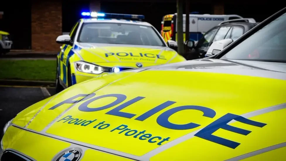 Two bright yellow police cars. 'Police - Proud to Protect' is written on both bonnets in blue letters. The blue lights are flashing on one of the vehicles.