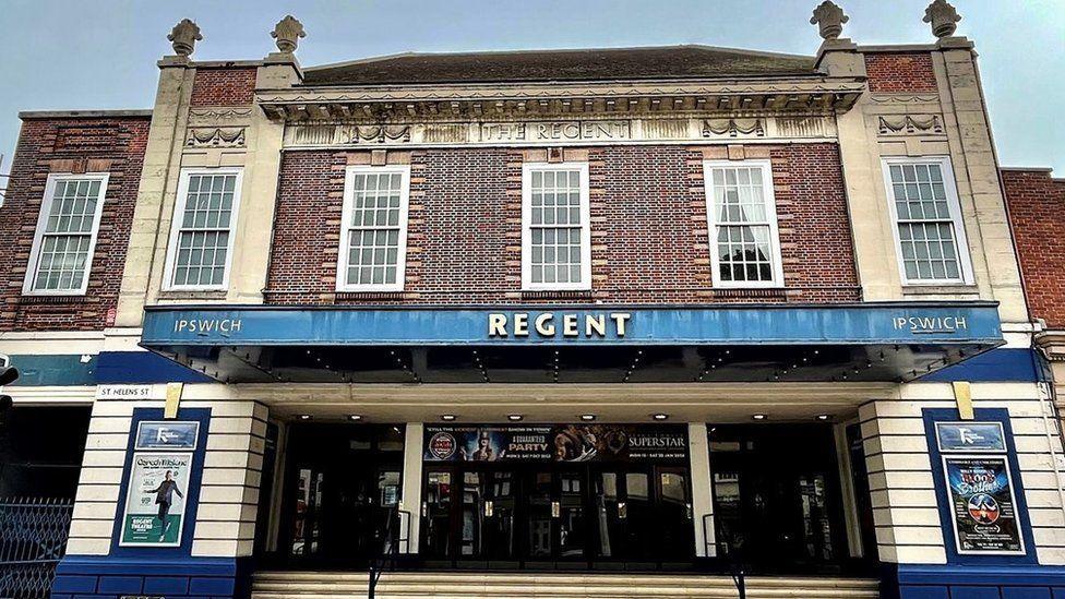 The exterior view of the Regent Theatre in Ipswich. The theatre name can be seen above the large entrance with multiple doors leading inside. Windows are above the entrance. Show posters have been placed either side of the entrance on the building.