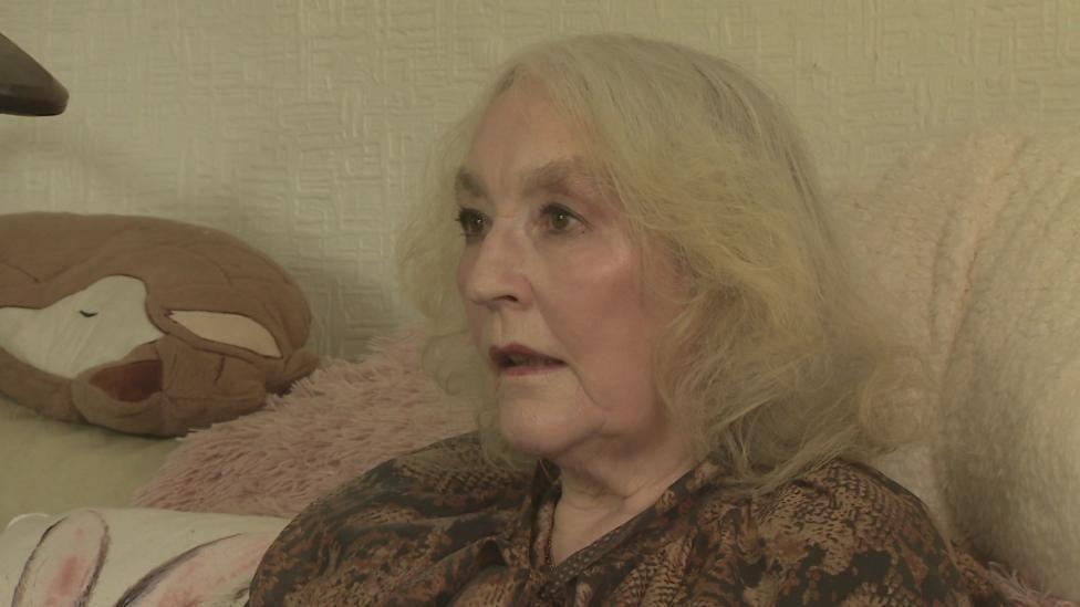 A woman with white hair in the style of bob sits on a sofa in a living room. She has a blue cardigan on and a patterned scarf around her neck. 