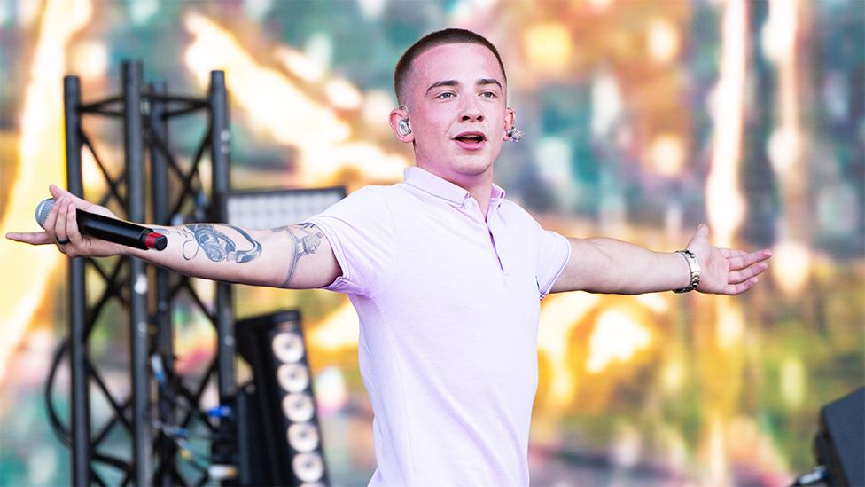 ArrDee looking at the crowd with his arms spread out with a mic in his right hand. he's wearing a light pink tshirt with a collar. He has an ear piece in his ears. Behind him is staging with multi coloured lights and metal railing holding up light and sound equipment.