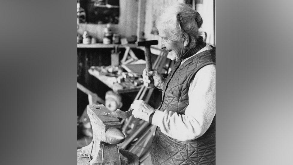 An elderly woman holding a hammer, hammering a piece of metal on an anvil