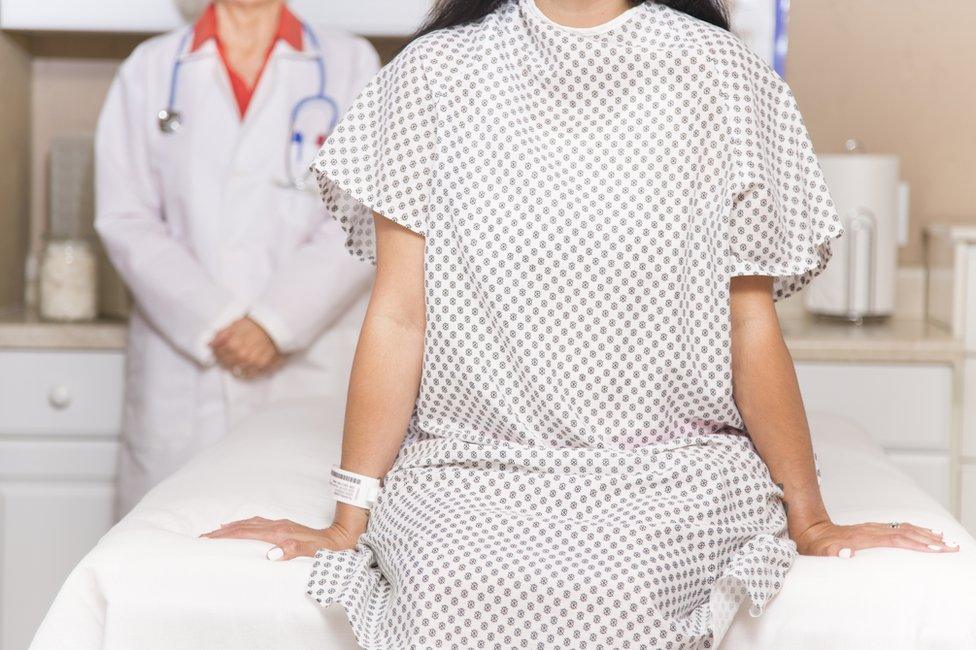 Female patient sitting on a trolley and wearing a hospital gown with a doctor standing behind her