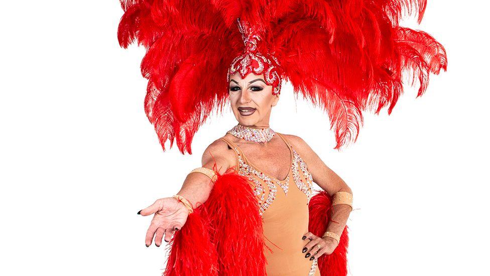 Zoe in full drag wearing a red showgirl outfit with a huge flamboyant red feather headdress, with her left hand on her hip and he right hand extended, standing against a white background