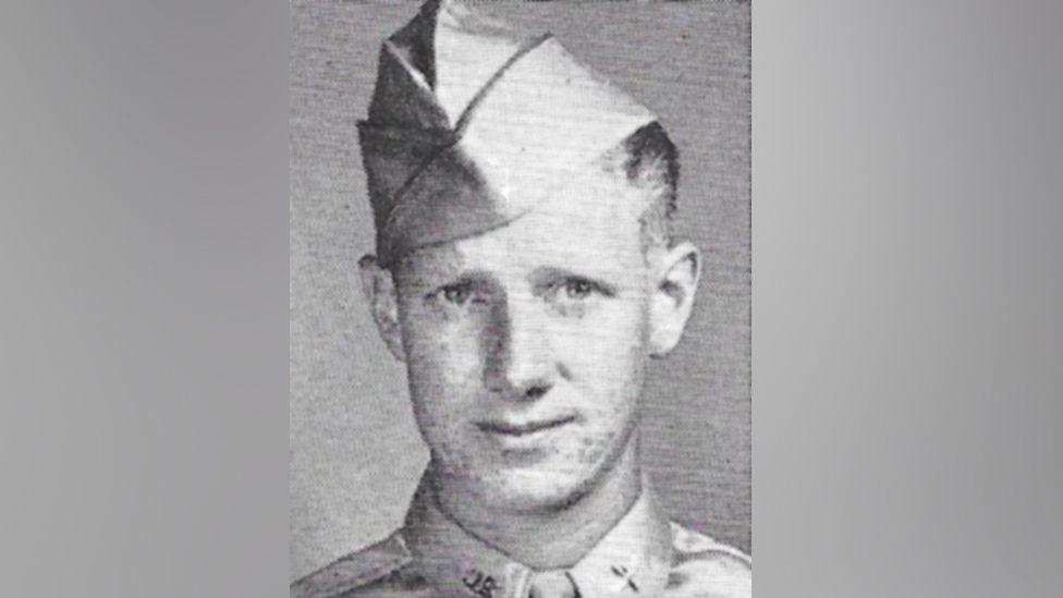 A grainy black and white image shows a young man in military uniform looking at the camera. Only his head and shoulders can be seen. He is wearing a cap and a shirt and tie