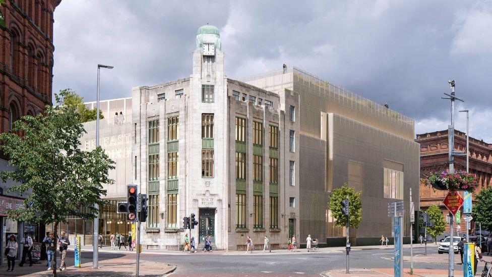 Image of the former bank of Ireland in Belfast. It is a tall white art deco building with green detailing standing at a street corner in front of a junction. Traffic, including buses and cars, moves along the road beside rows of red‑brick and modern buildings under a bright, partly cloudy sky.
Computer rendering shows how the extended Belfast Stories building will appear.
