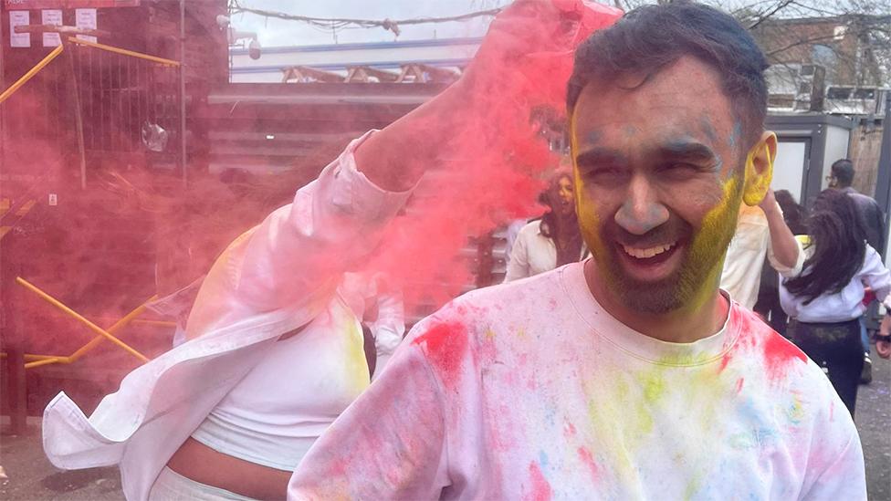 Almir, a man, wearing a white tshirt which has red and yellow powdered colour on it. He is smiling, with his beard coloured yellow and someone behind him throwing red powdered colour on him as they celebrate.