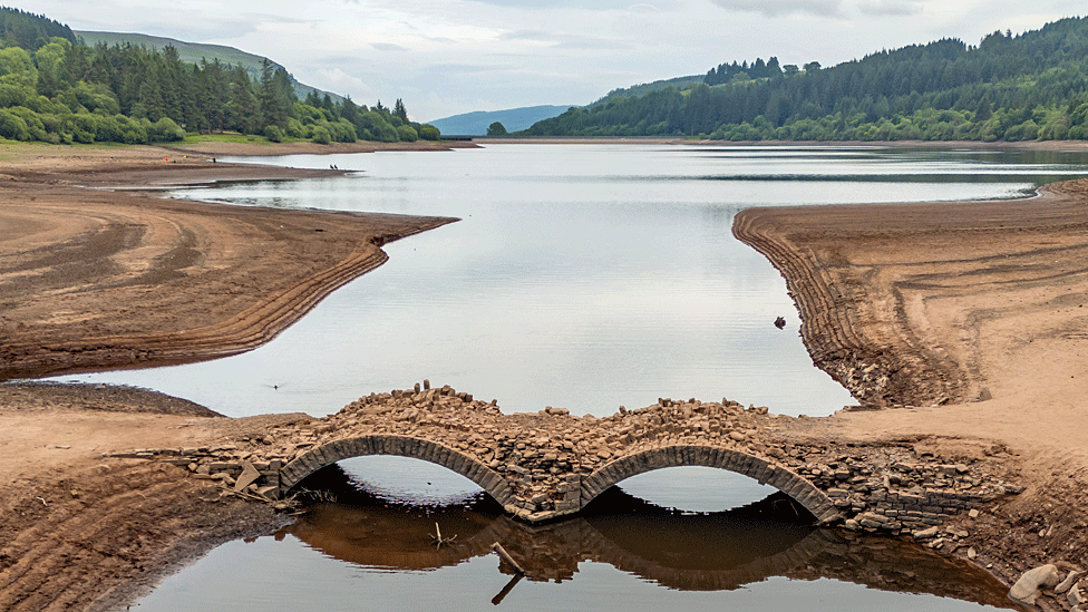 Two-arch stone bridge emerges in a largely dry bed forming part of today's reservoir