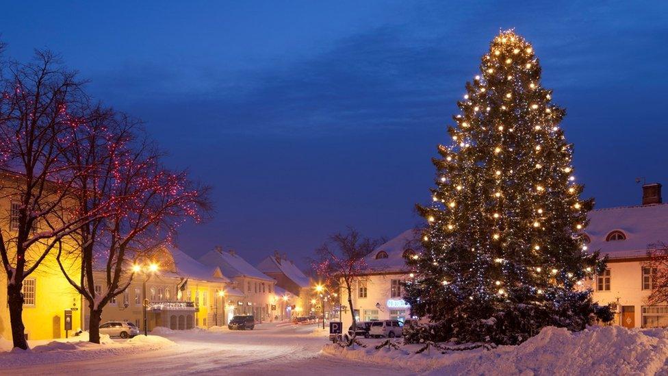 A lit Christmas tree on the right sits beside a road covered with snow with houses on either side. There is a tree with red lights on the left and the sky is deep blue.